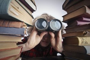 Woman looking through binoculars crouched between two stacks of books.