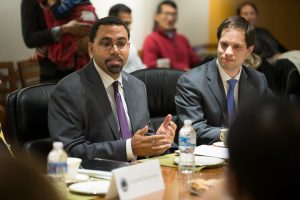 Two men siting at a table discussing meeting topic with others looking on from a row of chairs behind them.